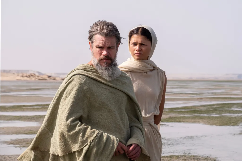 Matt Damon as Odysseus and Zendaya as Athena standing in a barren landscape.
