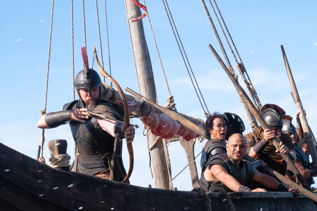 Matt Damon as Odysseus in “The Odyssey” film, aiming a bow and arrow from a boat with other warriors.