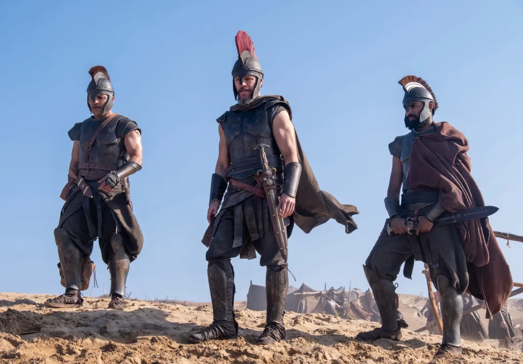Jimmy Gonzales, Matt Damon, and Himesh Patel in costumes as Cepheus, Odysseus, and Eurylochus, respectively, stand in a desert.