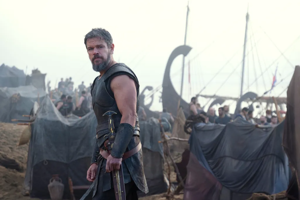 Matt Damon as Odysseus in warrior attire with a sword at his side, standing amidst a campsite with ships in the background.