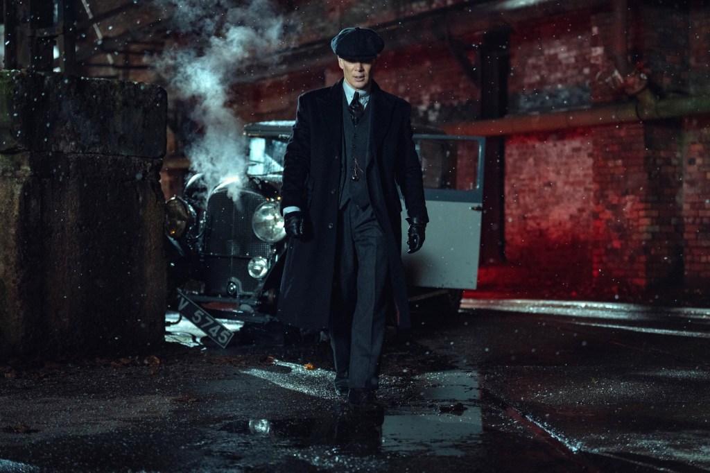 Cillian Murphy walking away from a car with an open door, smoke, and falling snow.