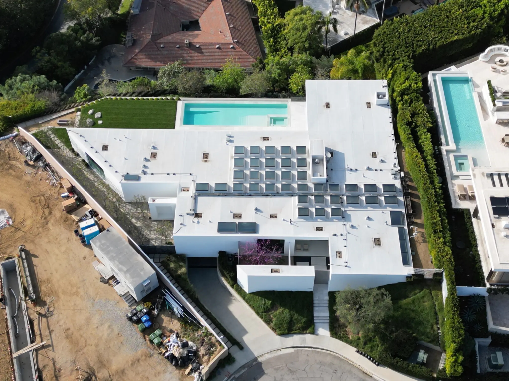 An aerial view of the Hollywood Hills home designed by XTEN Architects, featuring a white flat roof, multiple skylights, and a long swimming pool, with an adjacent construction site.