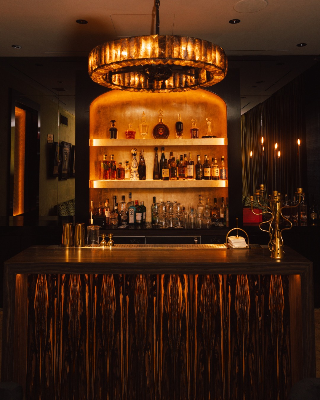An opulent bar at Zero Bond with a golden chandelier, illuminated shelves of liquor bottles, and a patterned wooden bar counter.