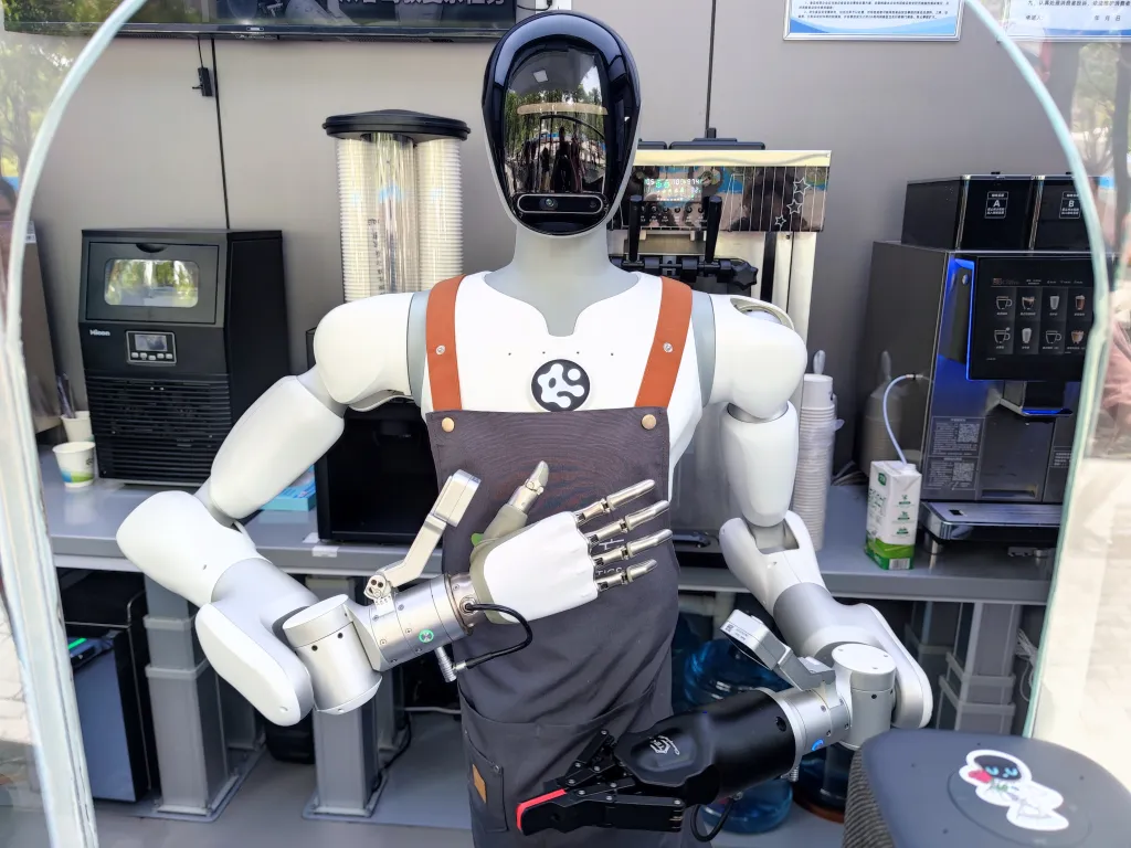 A robot barista in a gray apron stands in front of various coffee-making machines.