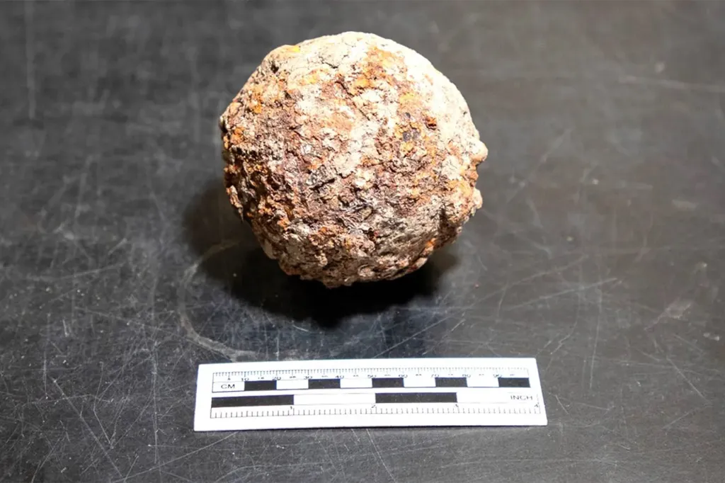 An ancient, rusted cannonball rests on a dark surface next to a ruler for scale.