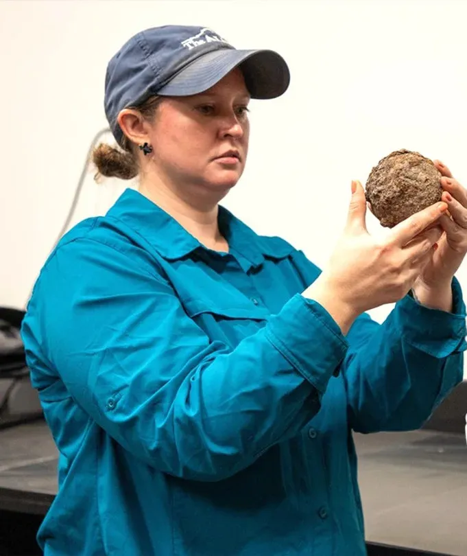 Woman in a teal shirt and hat holding an iron cannonball.
