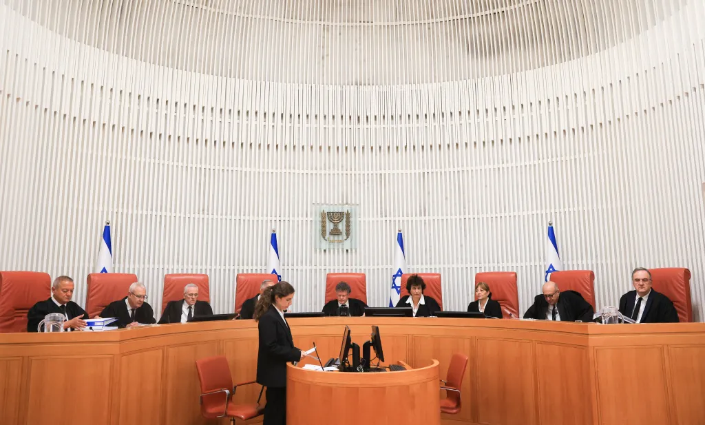 Israeli Supreme Court judges sit at a long curved wooden bench, with Israeli flags and the state emblem behind them.