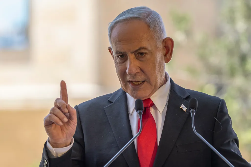Benjamin Netanyahu speaking at a podium with two microphones, wearing a suit, red tie, and an Israeli flag lapel pin, raising his right hand with his index finger pointed up.