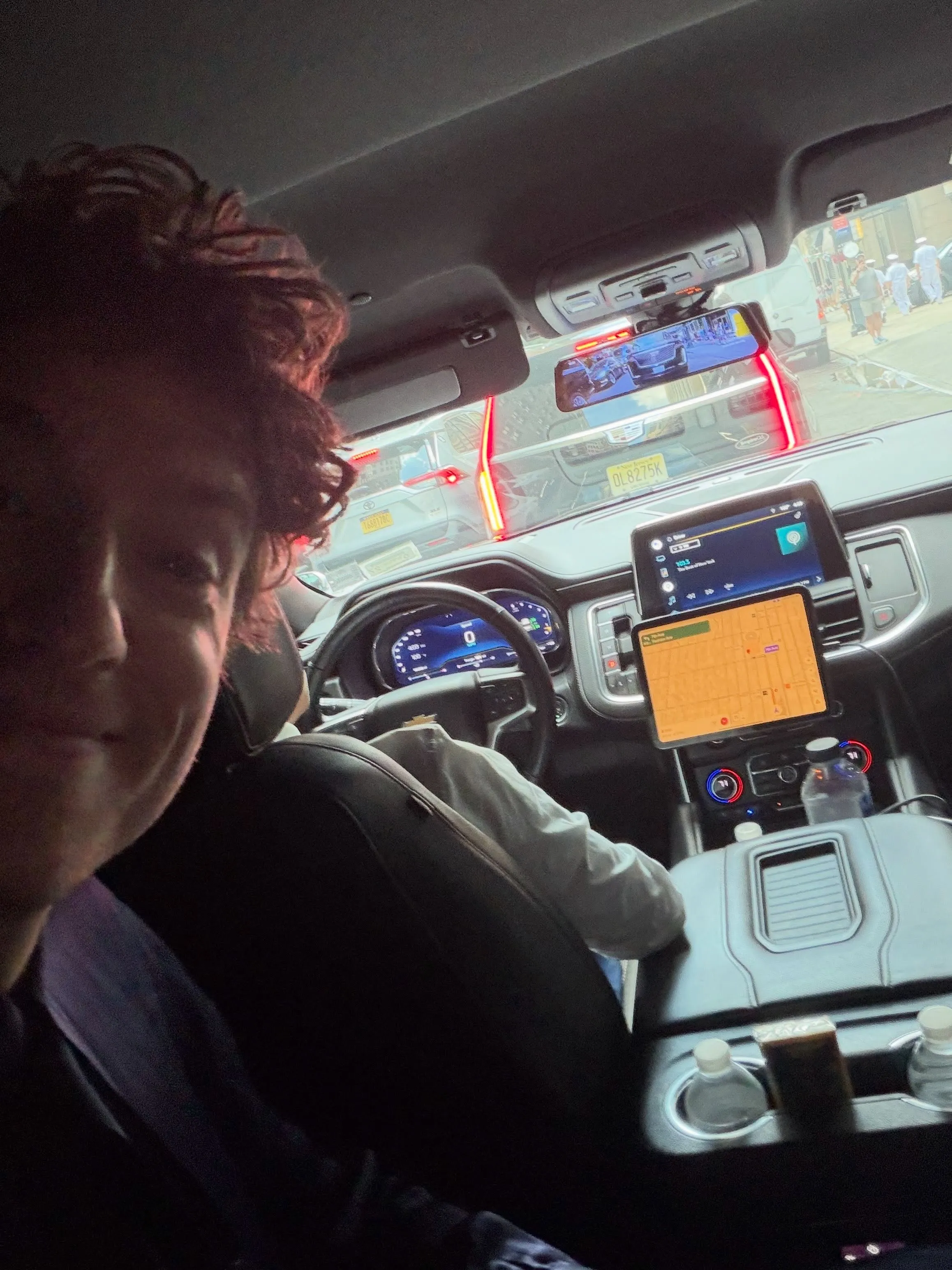 A person taking a selfie inside a car, with a street view of cars and people on the road in the background, visible through the windshield and rearview mirror.