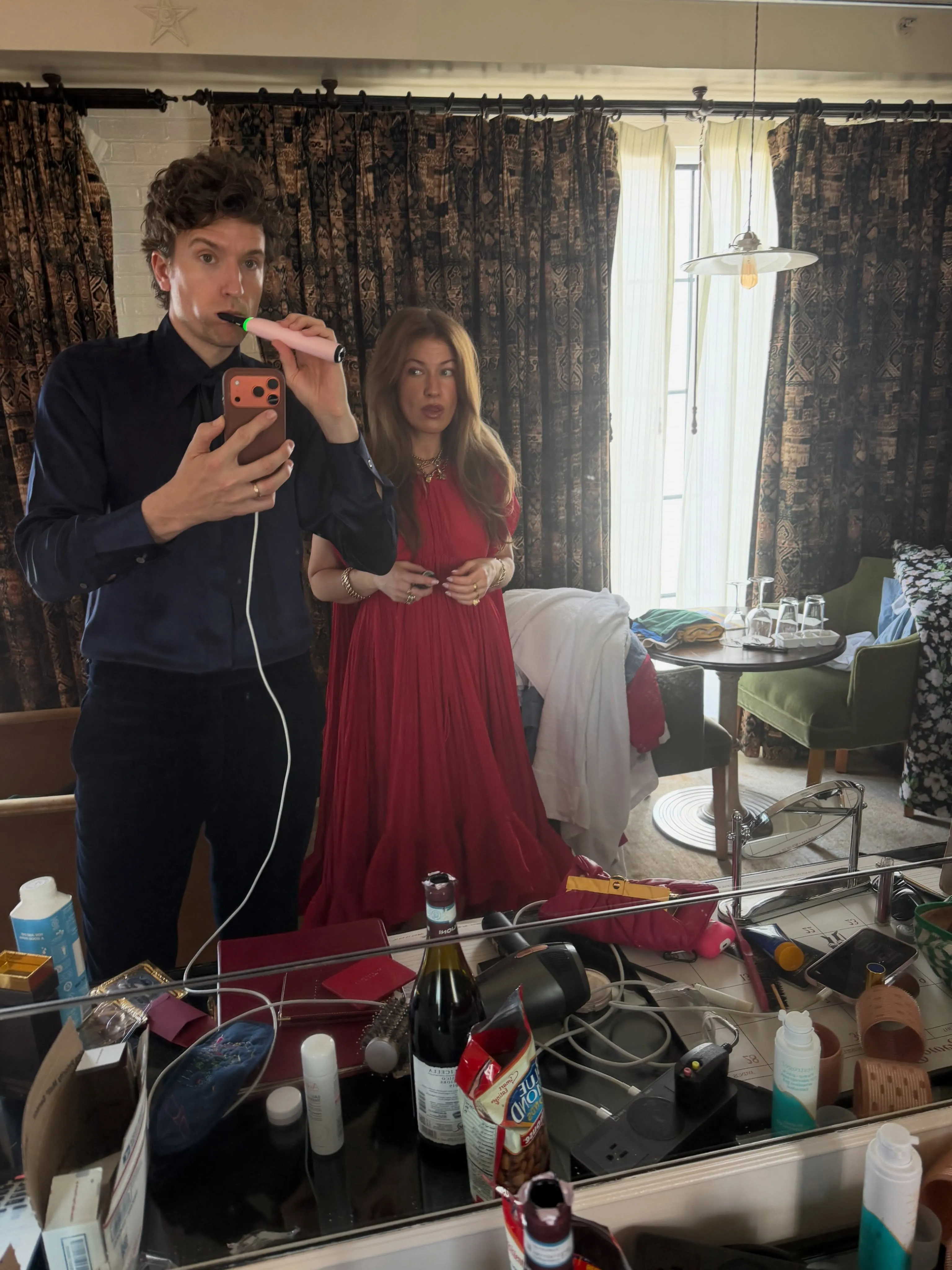Man brushing his teeth in a messy hotel room mirror with a woman in a red dress behind him.
