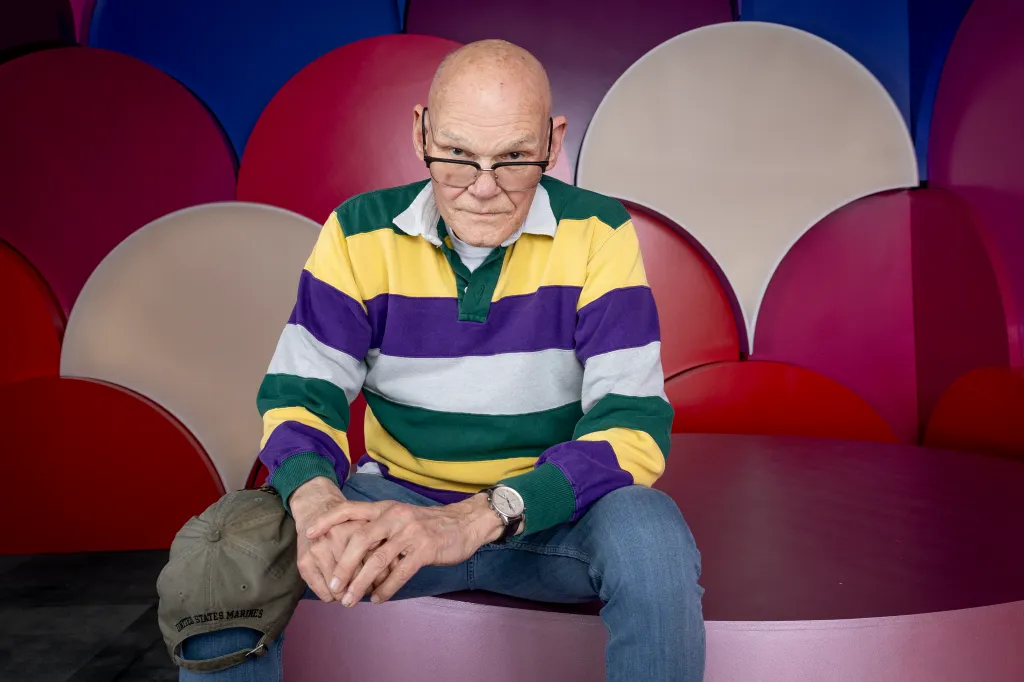James Carville in a yellow, purple, white, and green striped shirt, looking seriously at the camera with glasses, against a background of colorful half-circles.