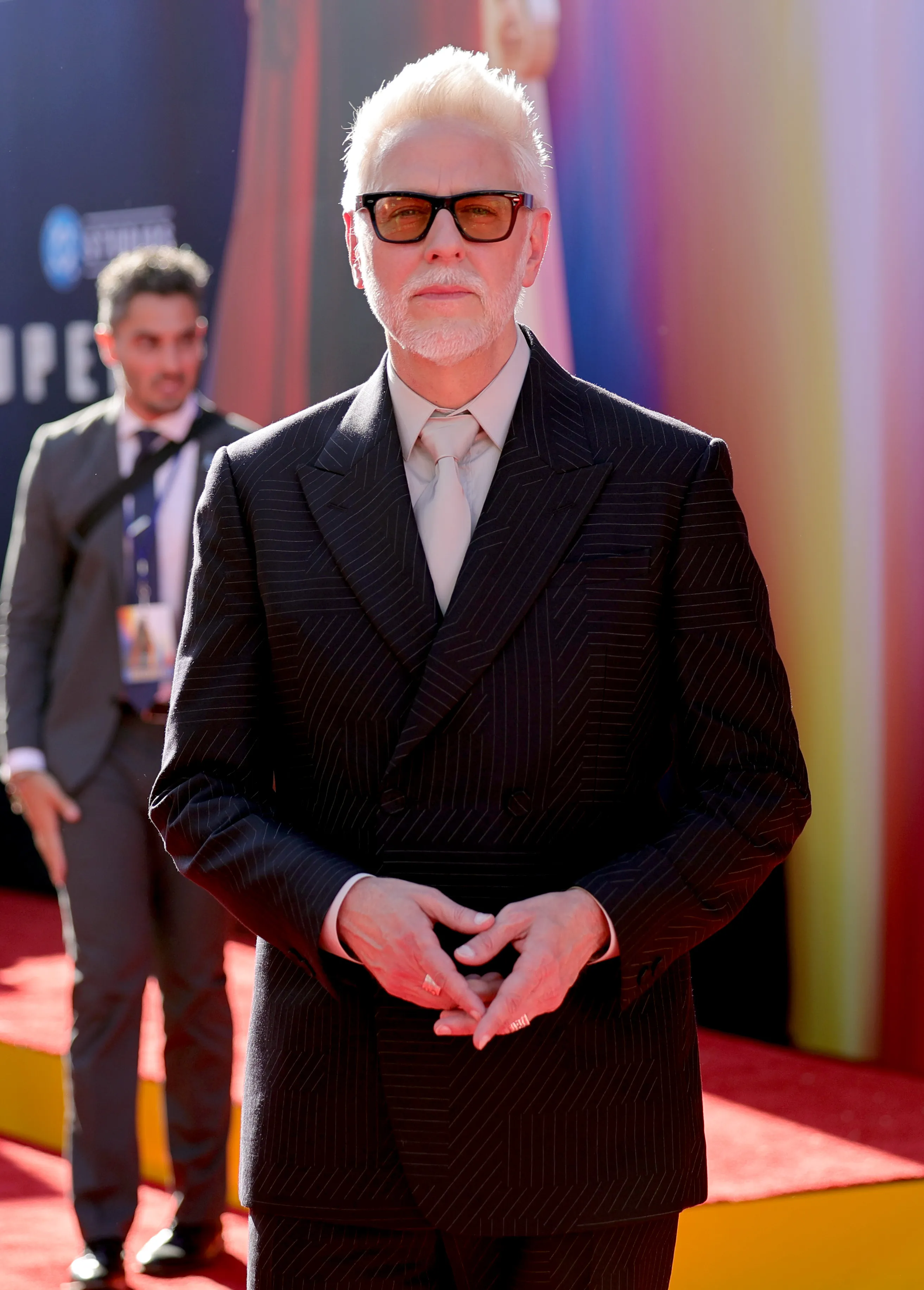 James Gunn, Co-Chairman and CEO of DC Studios, wearing a dark pinstripe suit, light tie, and sunglasses on a red carpet.