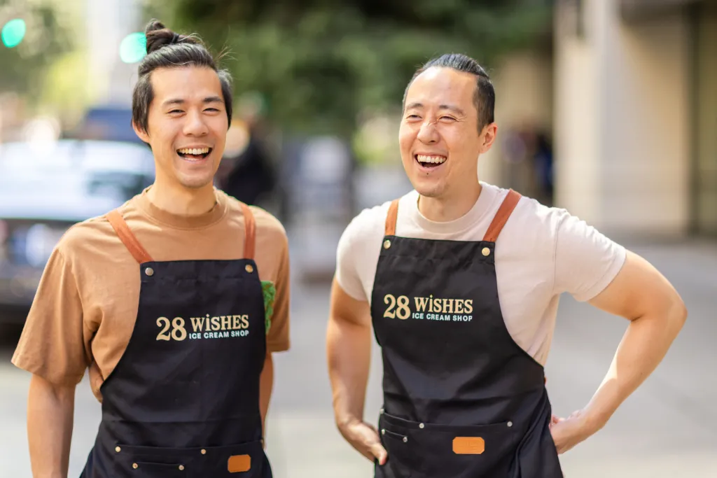 Brothers James and Jason Jiang, wearing black aprons with "28 Wishes Ice Cream Shop" printed on them, laugh while posing for a portrait.