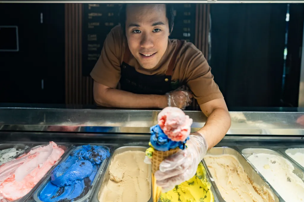 James Jiangon, wearing an apron and clear gloves, offers a multi-scoop ice cream cone from behind a display case with various ice cream flavors.