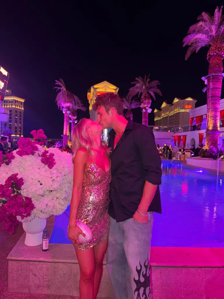 James Kennedy and Jordan Meyers kissing at night by a pool with palm trees, with Caesar's Palace in the background.