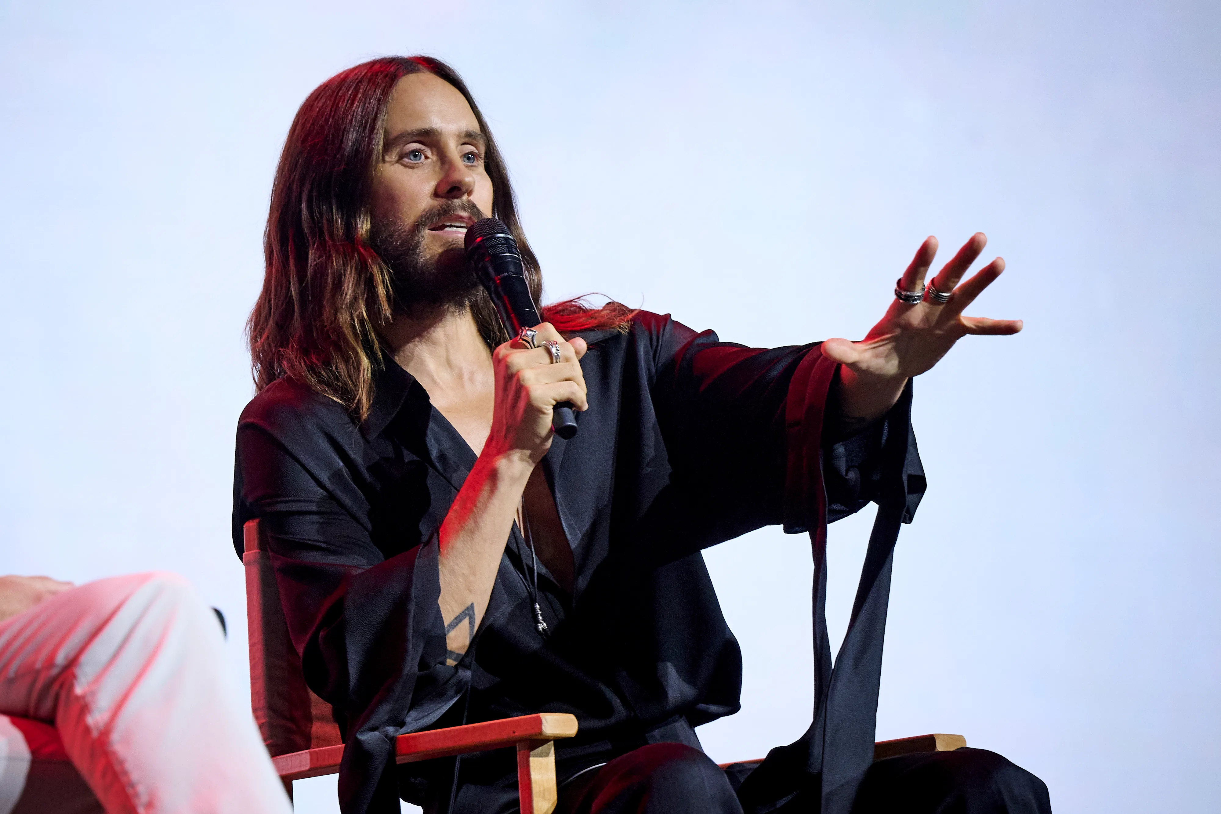 Jared Leto speaking into a microphone, wearing a black shirt and rings, with his arm outstretched at San Diego Comic-Con Malaga.