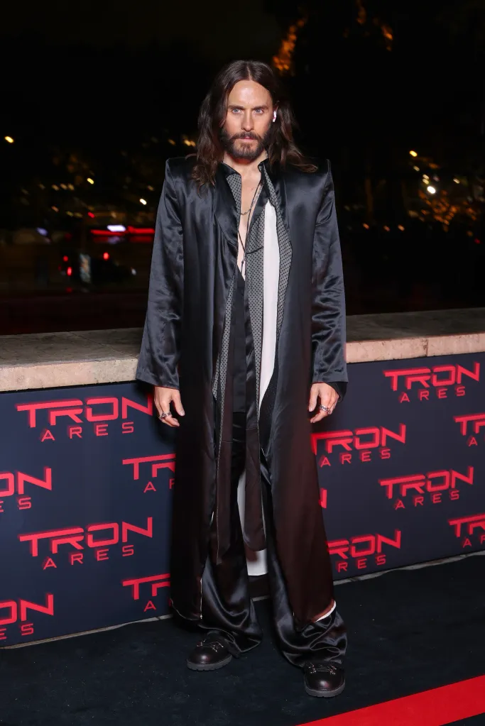 Jared Leto at the "Tron: Ares" Paris Photocall, wearing a long black coat over a patterned top, with long brown hair and a beard, standing on a red carpet with "TRON ARES" logos.