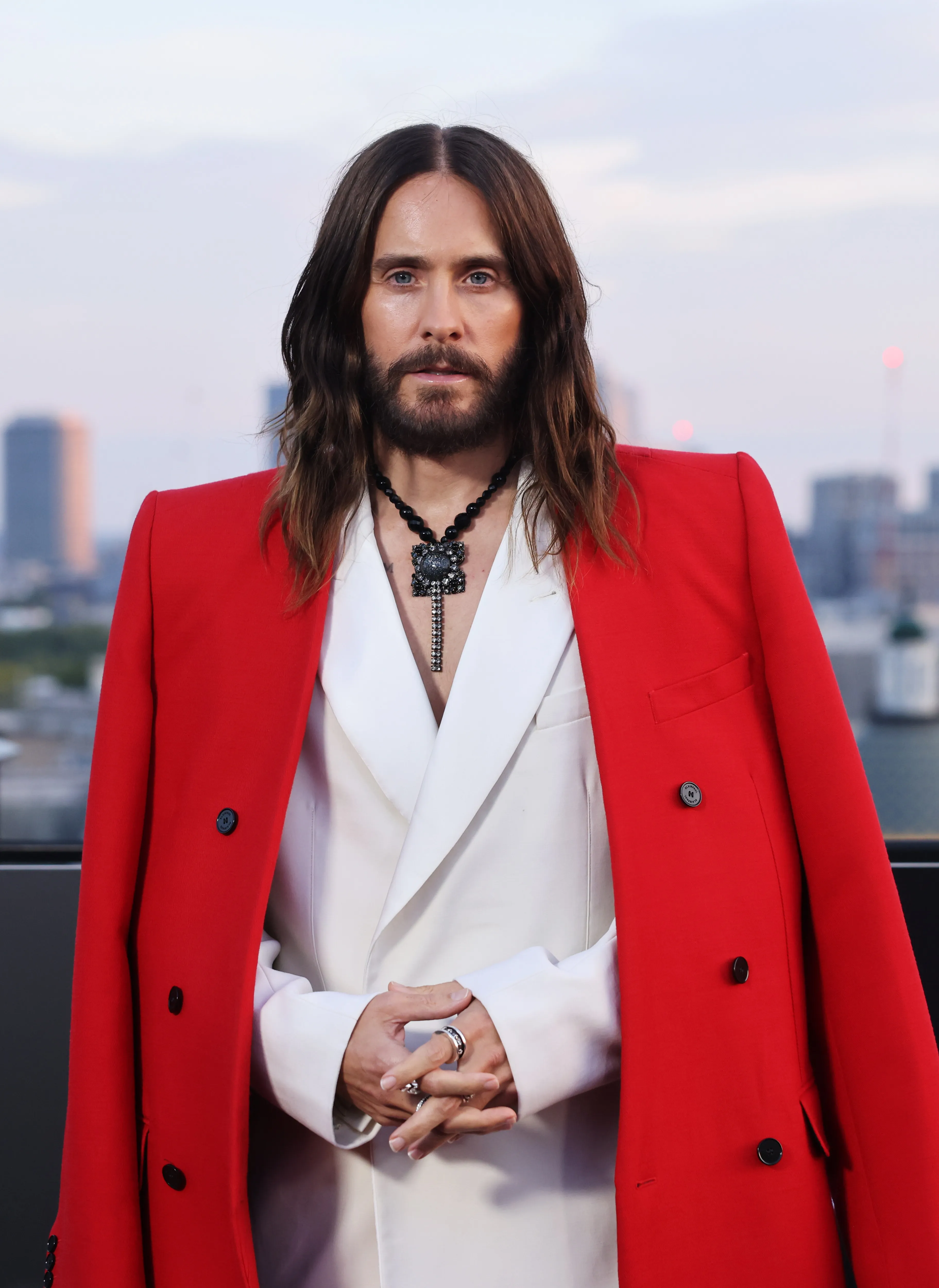Jared Leto posing for a photo in a white suit and red coat with the London skyline in the background.
