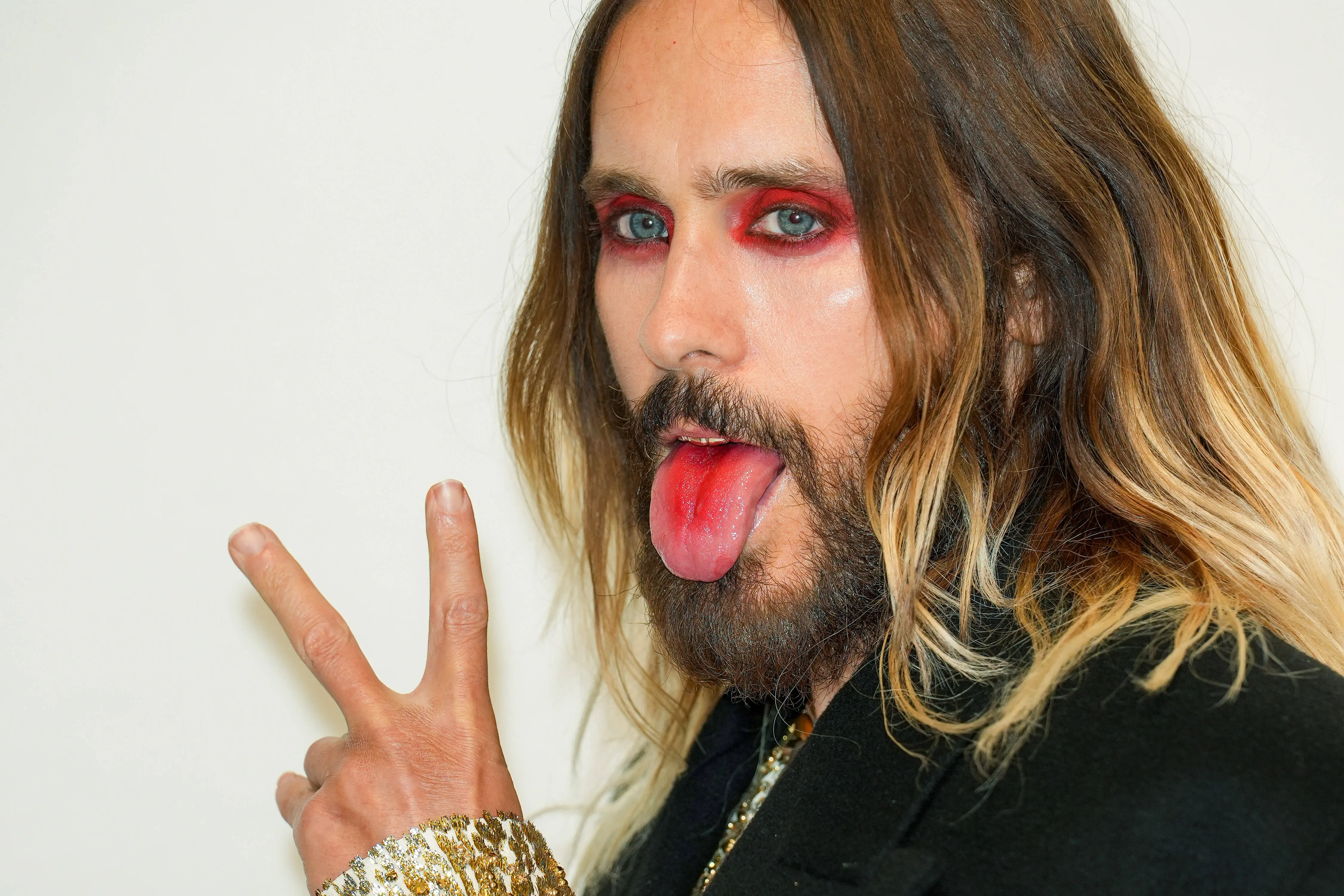 Jared Leto with red eye makeup, a beard, and long hair, sticking out his tongue and making a peace sign.
