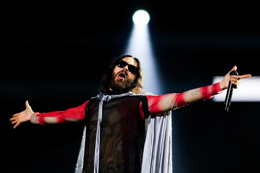 Jared Leto of Thirty Seconds to Mars performs on stage wearing sunglasses, a gray cape, and a sheer black and red top, with his arms outstretched and holding a microphone.