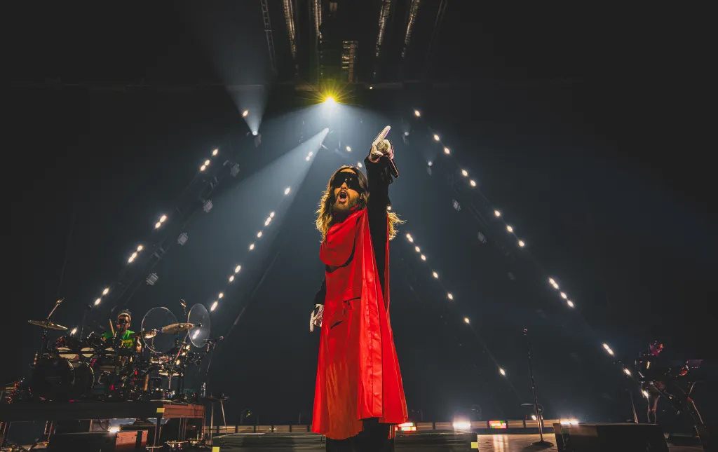 Jared Leto performing on stage in a red cape and sunglasses, raising a microphone in his gloved hand.