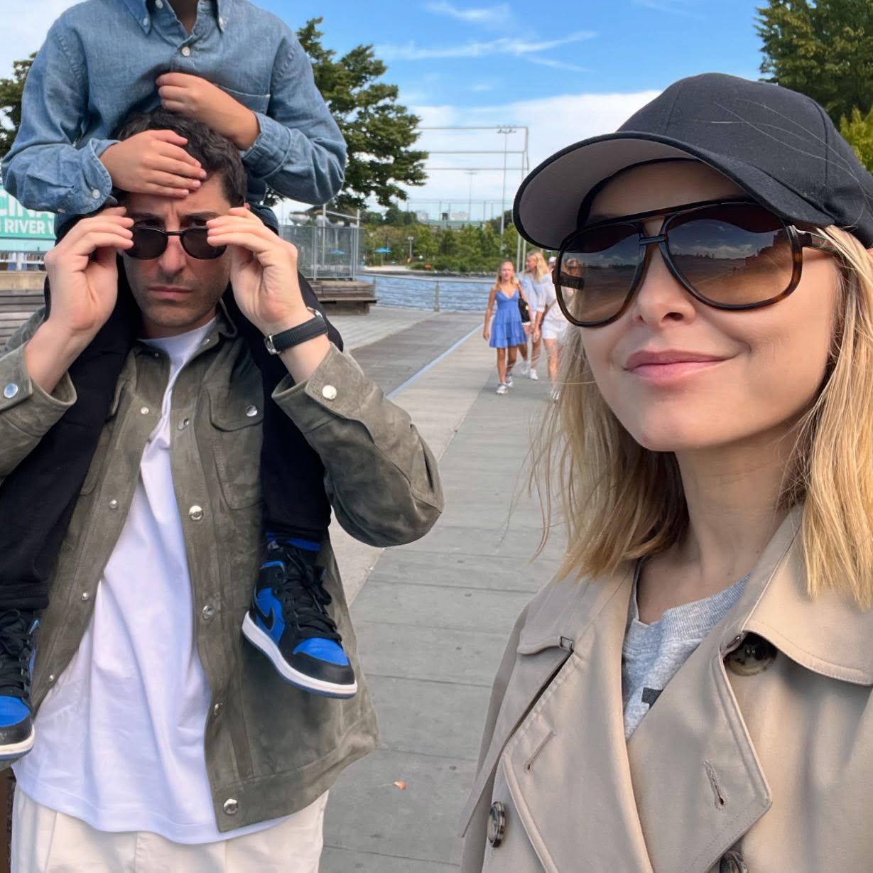 Jenny Mollen taking a selfie with Jason Biggs giving their child a piggyback ride by the water.