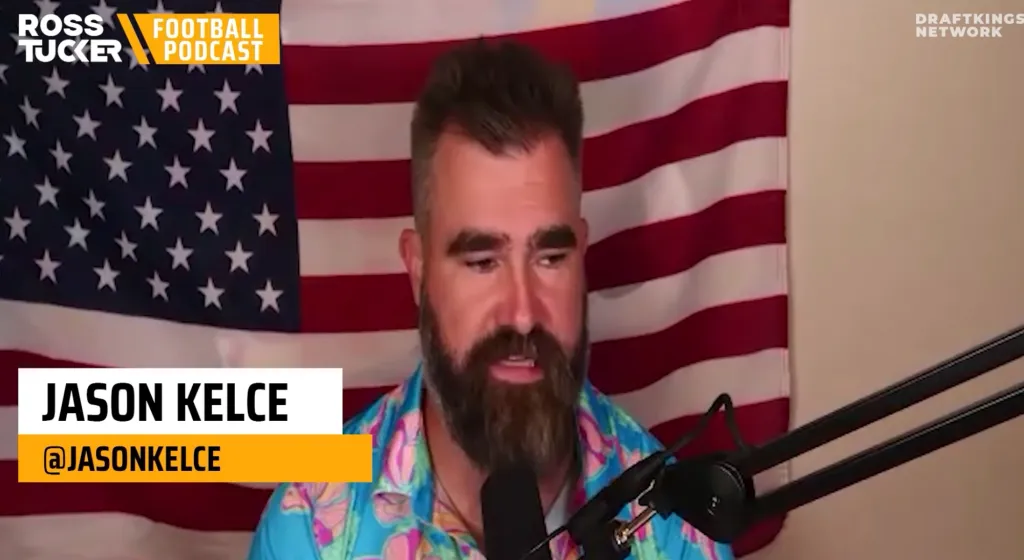 Jason Kelce speaks into a microphone in front of an American flag.