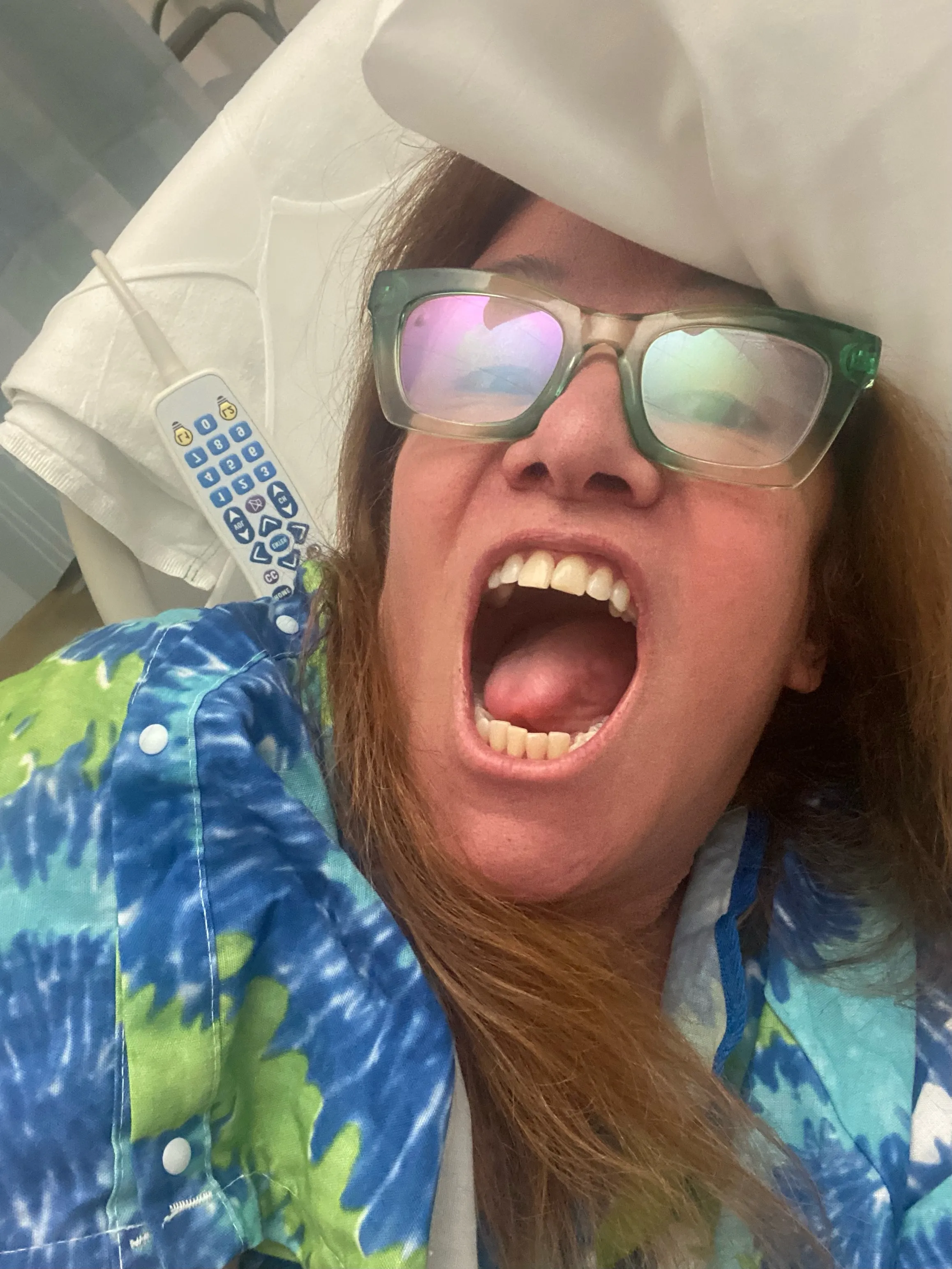 Jennifer Finch wearing a blue and green tie-dye shirt, green glasses, with her mouth wide open in a hospital bed.
