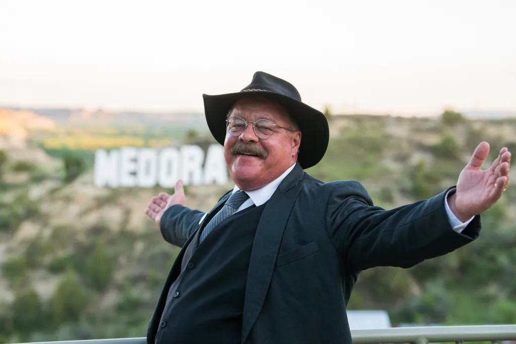 Joe Wiegand as Teddy Roosevelt with his arms outstretched in front of a "Medora" sign.
