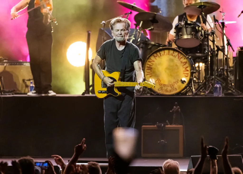 John Mellencamp playing an electric guitar on stage with a drum set behind him.