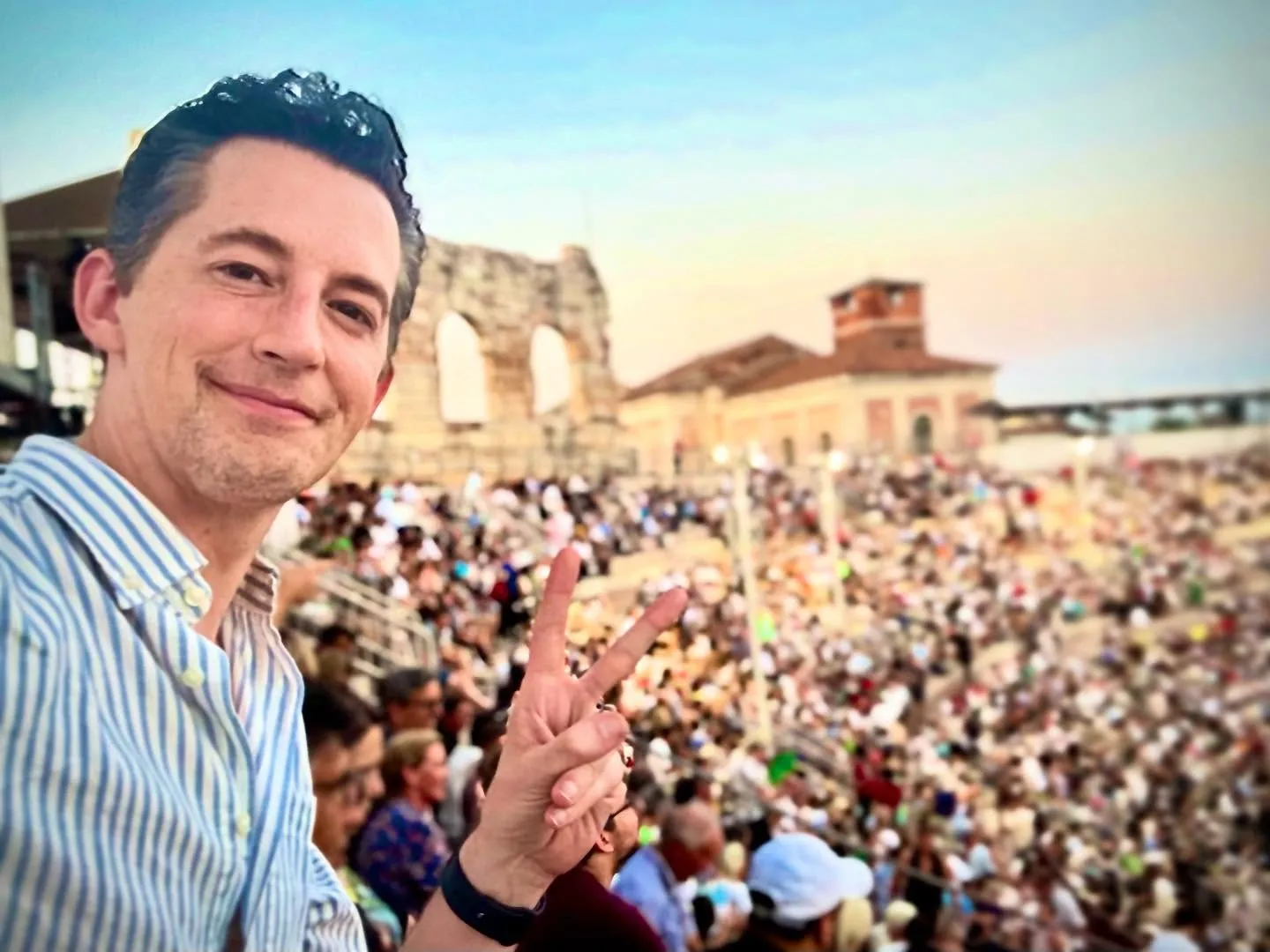 Josh Grisetti taking a selfie and giving a peace sign in front of a crowded amphitheater.