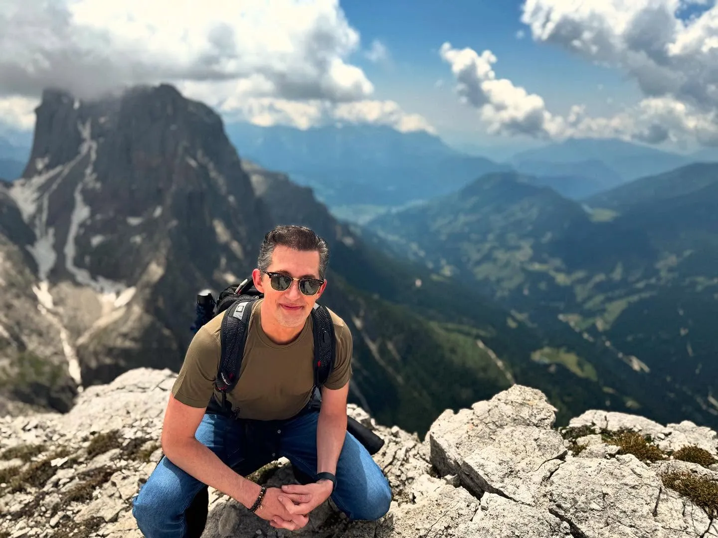 Josh Grisetti wearing sunglasses and a backpack, crouches on a rocky mountain peak with a scenic view of other mountains in the background.