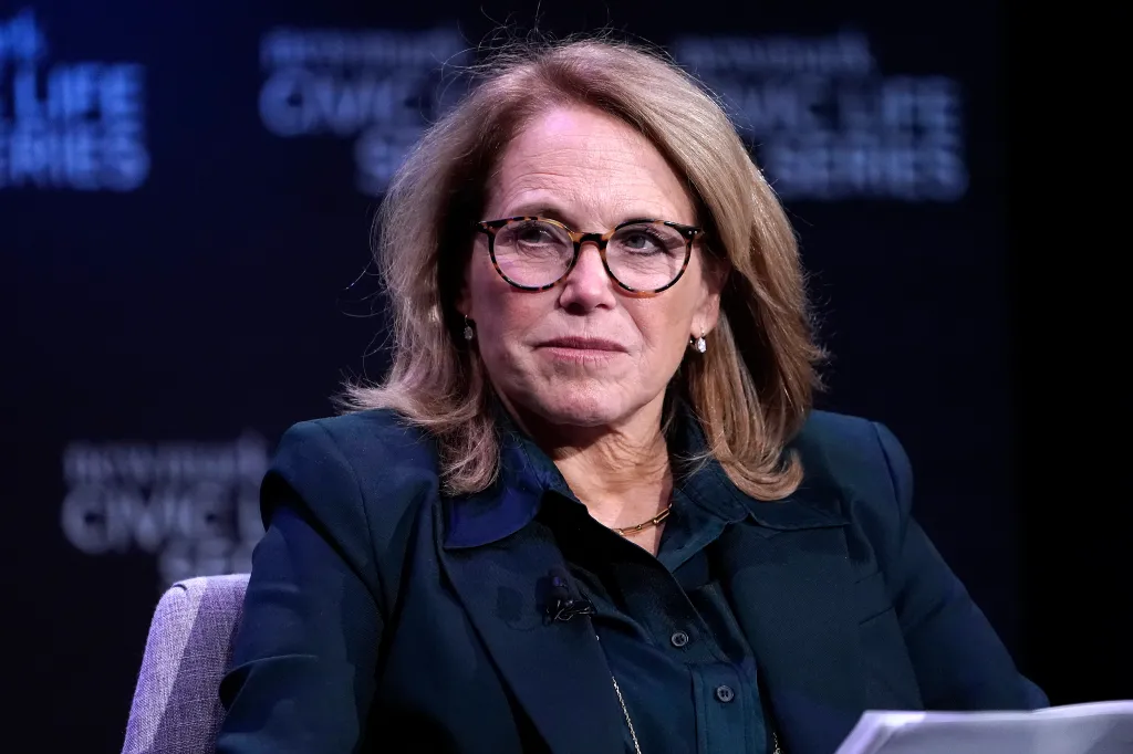 Journalist Katie Couric speaks at an event, wearing a dark green blazer over a dark collared shirt and tortoise-shell glasses.