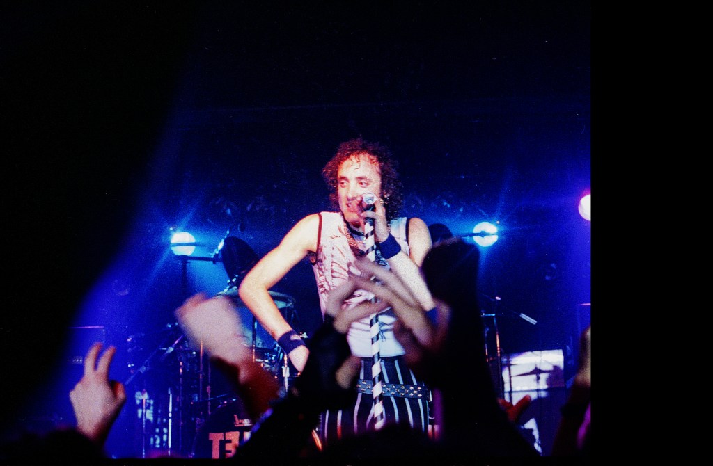 Kevin DuBrow of Quiet Riot performing on stage, bathed in blue light, holding a microphone.