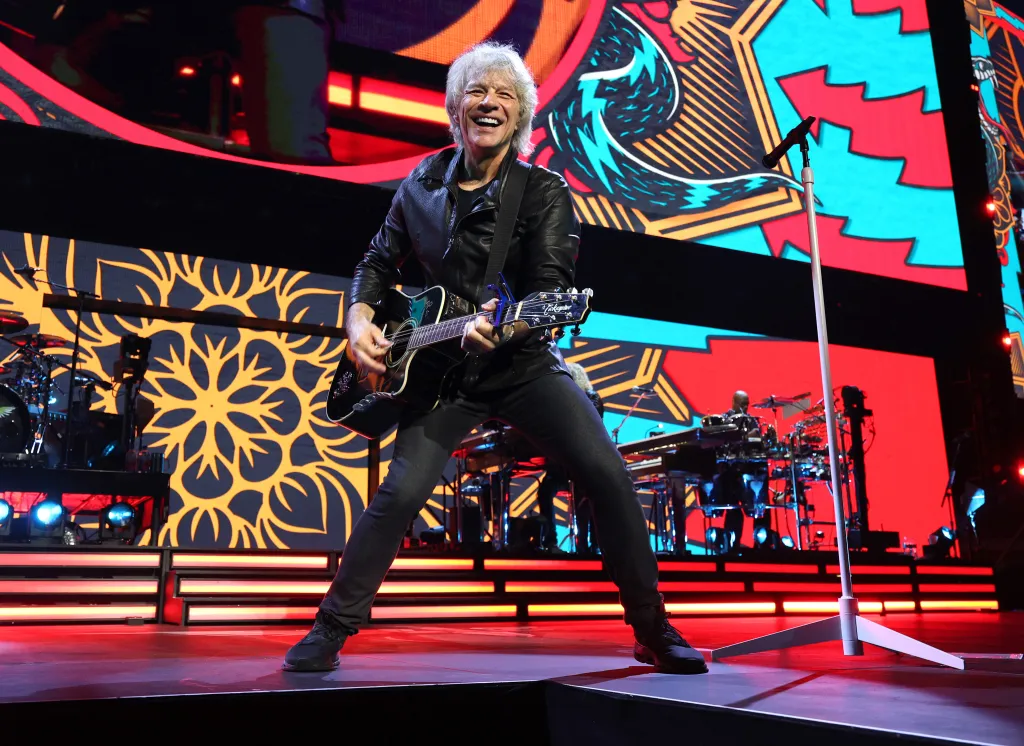 Jon Bon Jovi performing on stage during the Forever Tour, holding a guitar and smiling.
