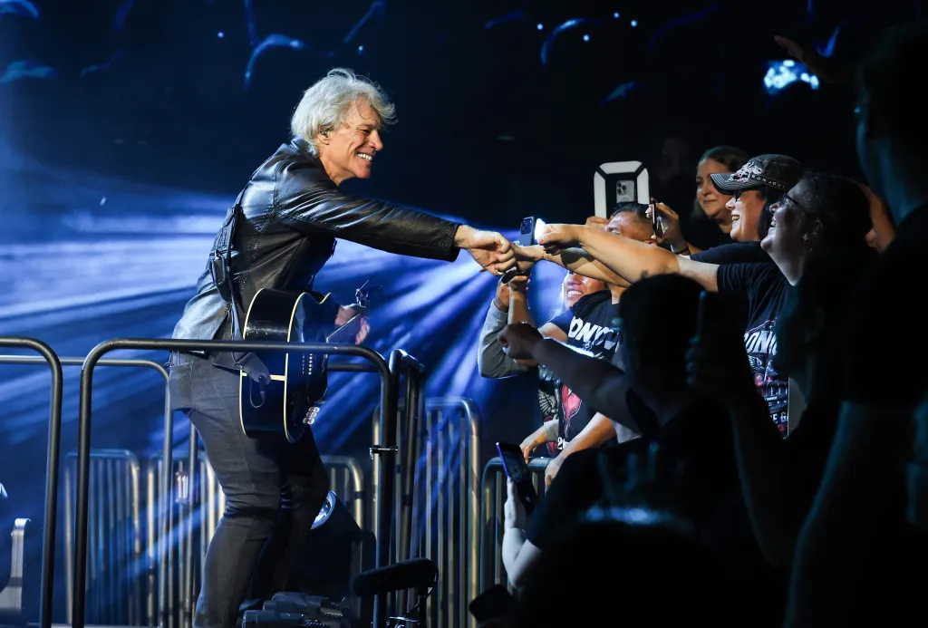 Jon Bon Jovi performing on the Forever Tour, leaning into the crowd to shake hands with fans while holding his guitar.