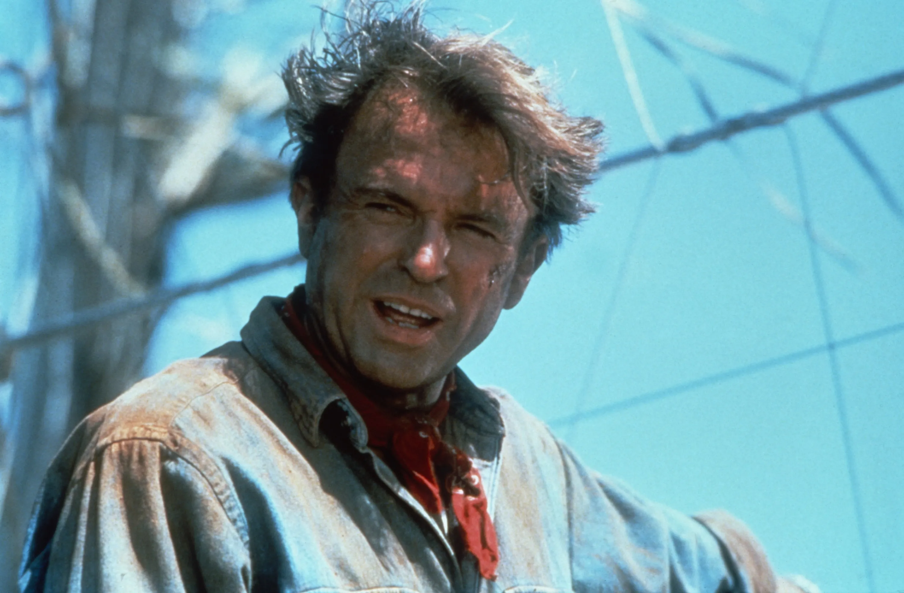 Portrait of Sam Neill as Alan Grant in Jurassic Park, with dirty, disheveled hair, wearing a denim shirt and red bandana.