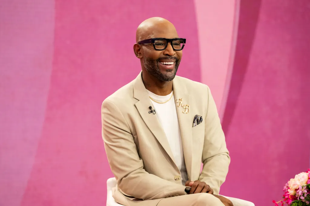 Talk show host Karamo Brown smiling and looking left, wearing a light tan suit and dark-rimmed glasses.