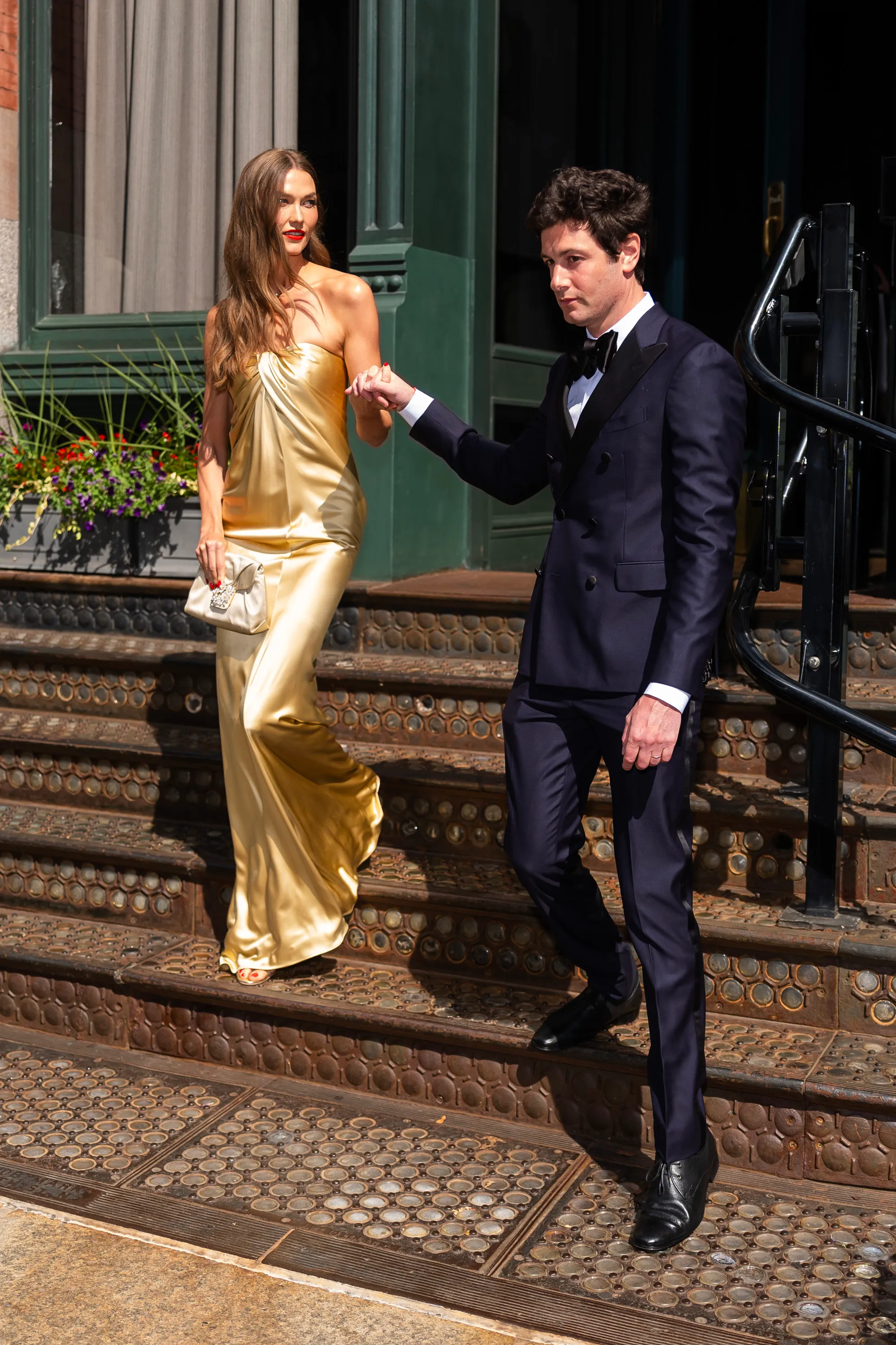 Karlie Kloss in a gold gown and Joshua Kushner in a navy tuxedo walk down stairs holding hands in SoHo.