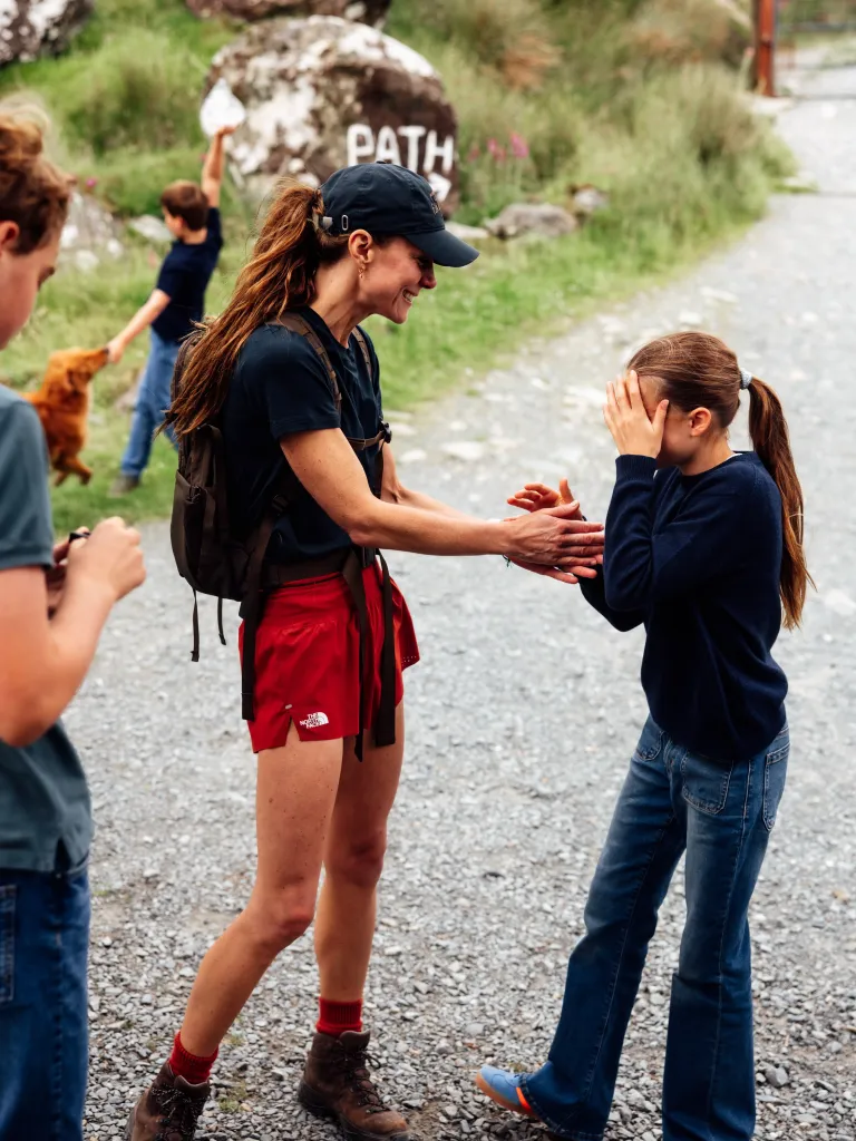 Kate Middleton wearing a dark cap, navy t-shirt, red shorts and hiking boots, holding hands with a child in a navy top and jeans, who is covering their face with their hands.
