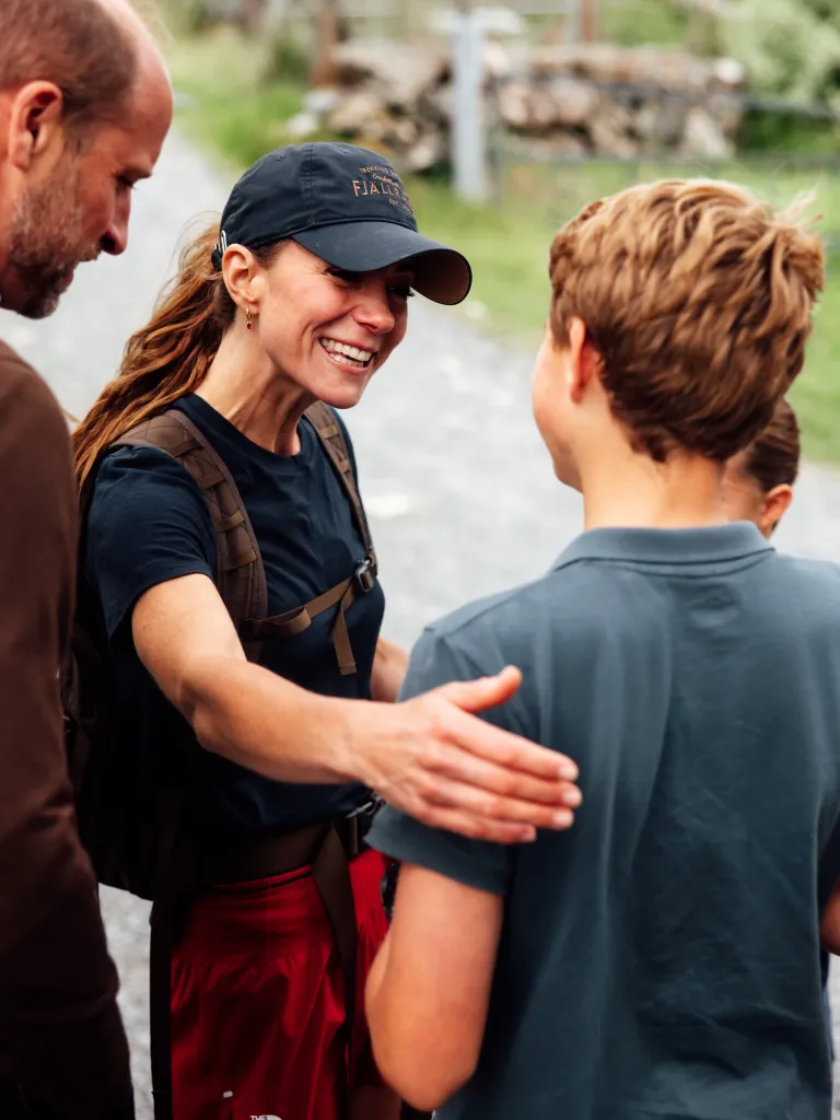 Kate Middleton smiling and wearing a baseball cap and backpack, with her arm around her son in a blue polo shirt.