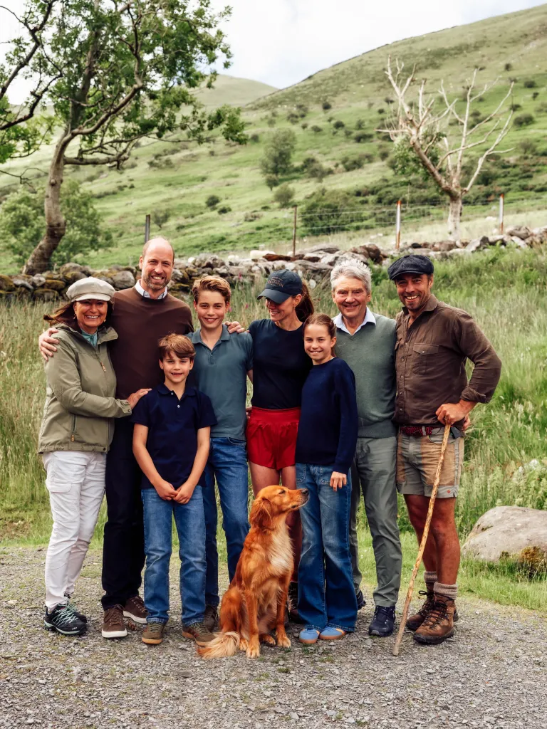 Kate Middleton posing with her three children and five other adults, with a dog at their feet, in a grassy, mountainous landscape.