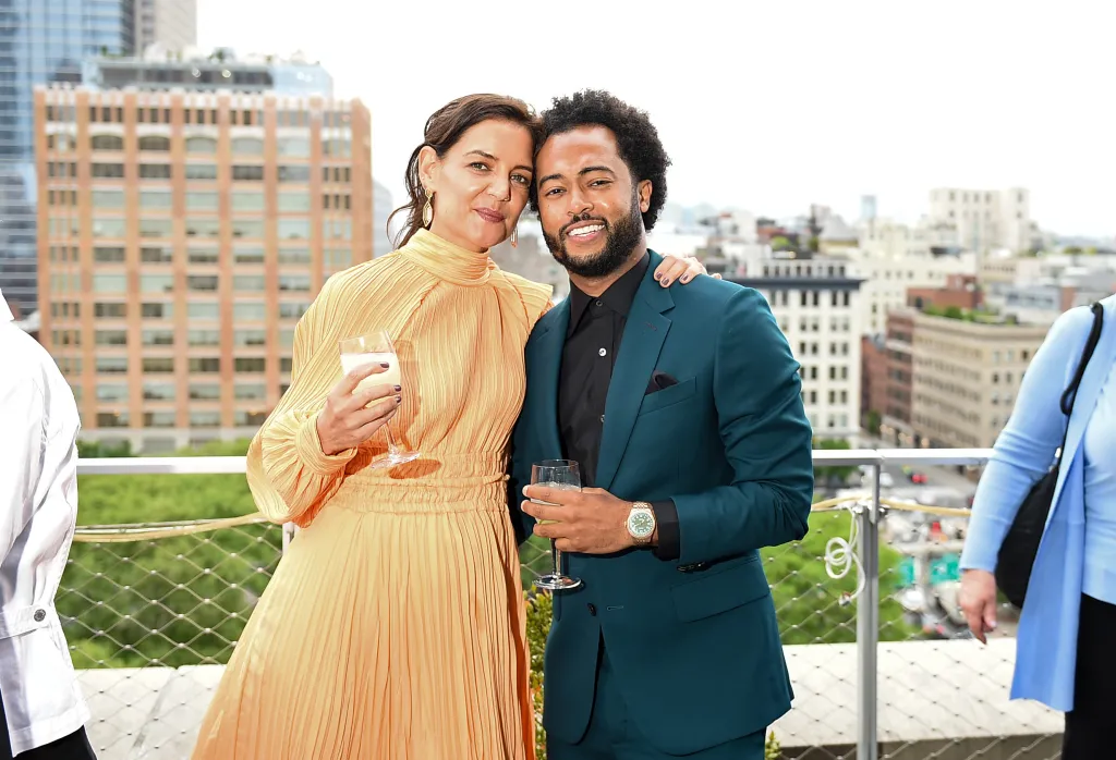 Katie Holmes in a gold dress and Bobby Wooten III in a dark teal suit pose on a rooftop.