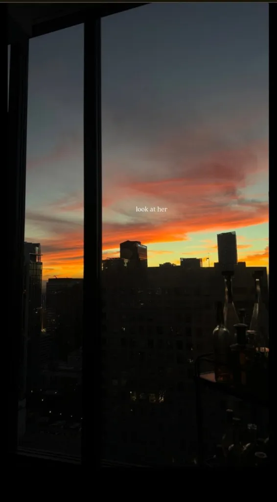 View of a vibrant sunset over a city skyline from a window, with the text "look at her".
