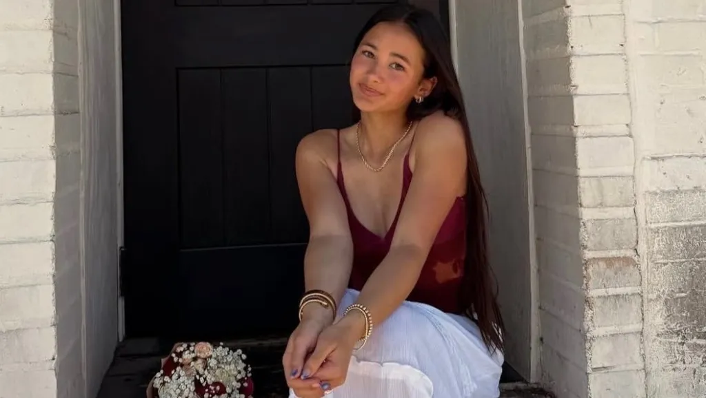 Kaylee Hottle, a young woman with long dark hair, wearing a red tank top and white skirt, sitting outside a doorway with a bouquet of flowers next to her.