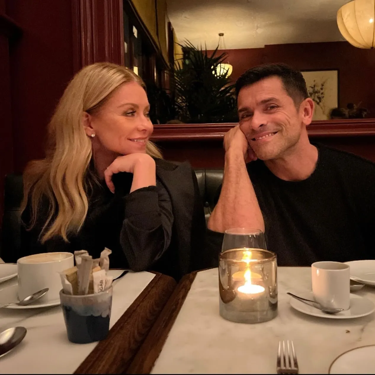 Kelly Ripa and Mark Consuelos looking at each other, sitting at a table with coffee cups and a lit candle.