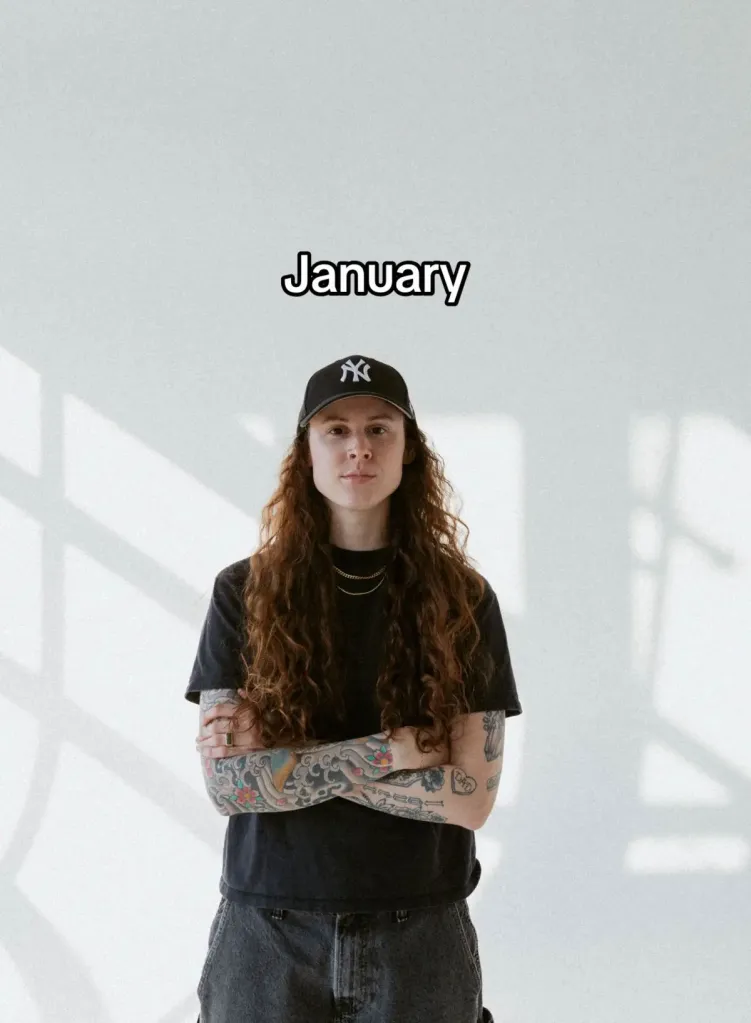Kelsie Chesser in a black New York Yankees cap with long curly hair, black t-shirt, and tattooed arms crossed.