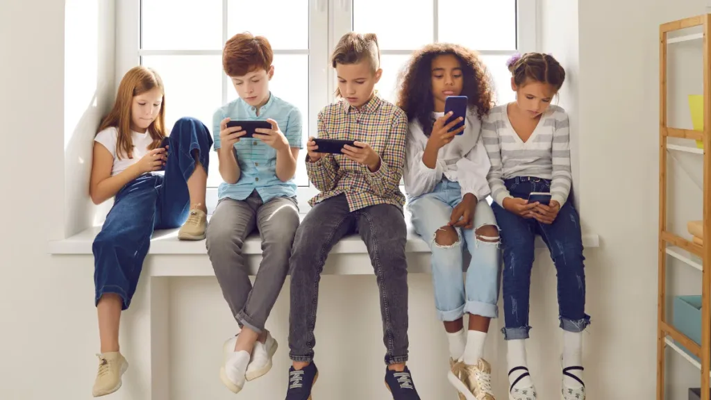 Group of children of different nationalities sit in a row on the windowsill and with lack of emotion play online games or read social networks on mobile phones.