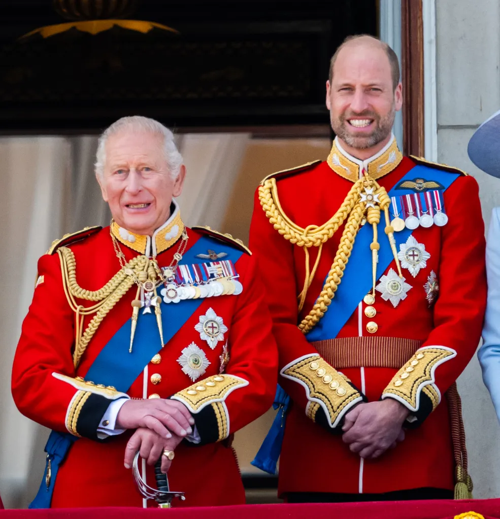 King Charles III and Prince William in red military uniforms with blue sashes and medals, standing on a balcony.