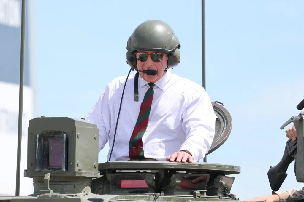 King Charles III in a tank helmet and sunglasses, speaking into a microphone while inside a tank.